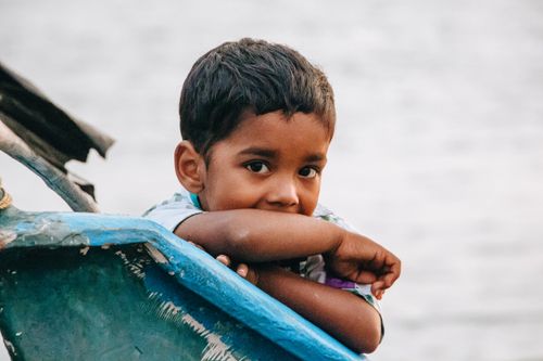 Free A contemplative child leans on a blue boat, gazing thoughtfully. The serene water backdrop adds tranquility. Stock Photo