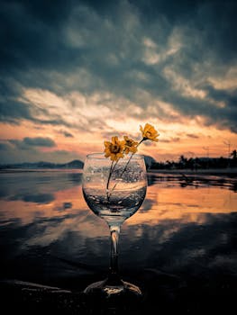 A serene wine glass with yellow flowers amidst a stunning sunrise reflection
