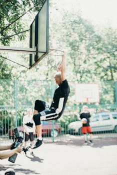 A young man hangs on a basketball hoop after a dunk on an outdoor court.