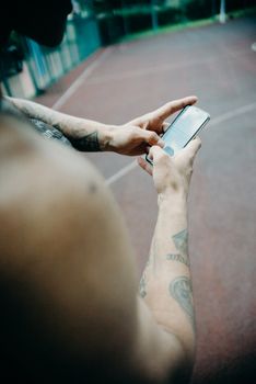 Tattooed athlete using smartphone on outdoor basketball court. Focus on mobile device and hand tattoos.