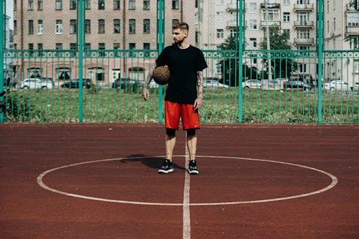 Athletic young man standing on an outdoor basketball court holding a ball, with urban buildings in the background.