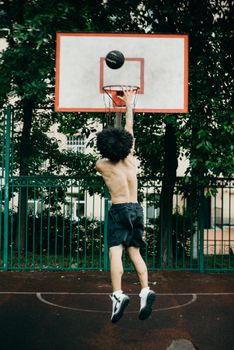 A shirtless man performs a dunk on a basketball court outdoors, showcasing athleticism and skill.