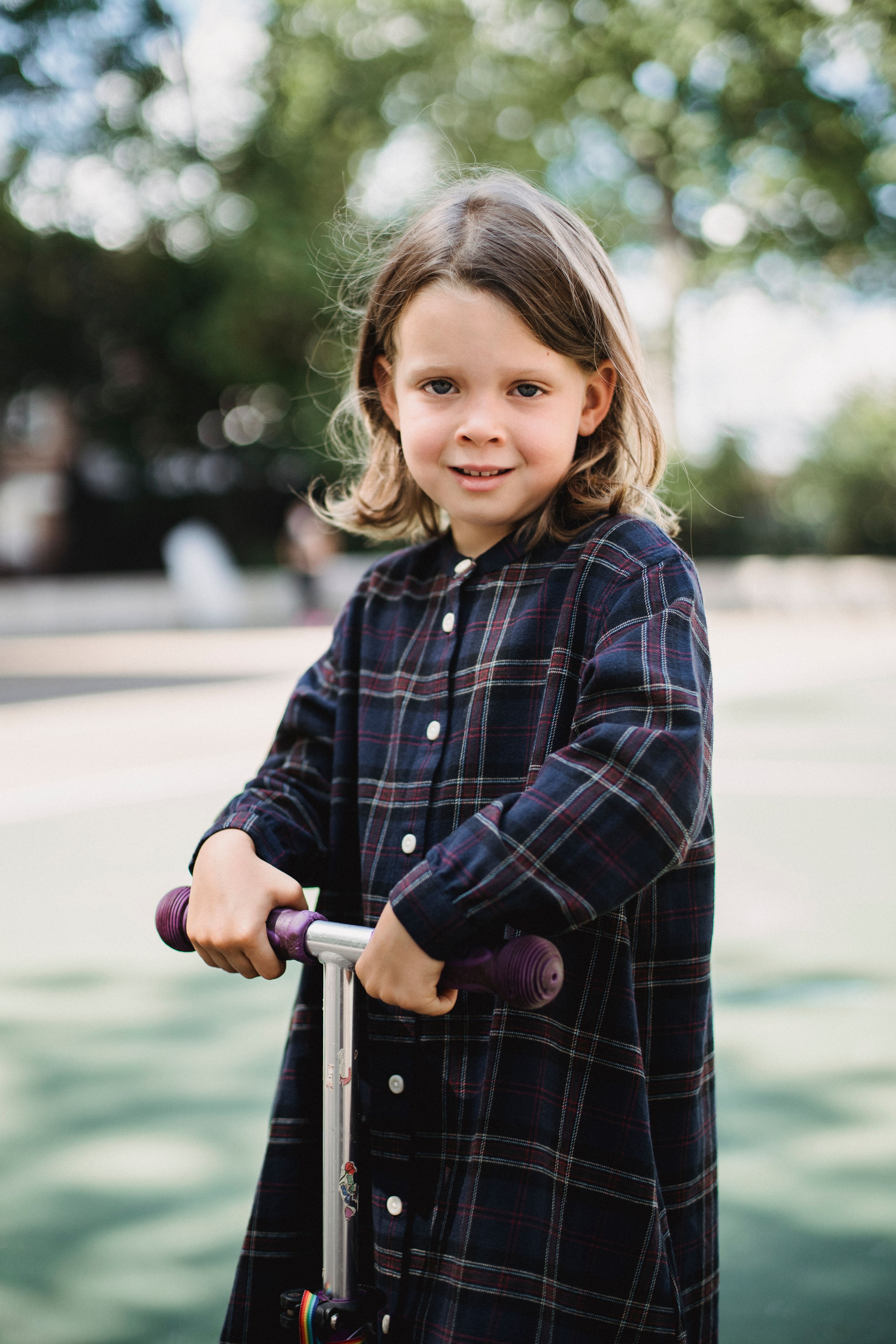 A smiling young girl with long hair stands joyfully on a scooter in a sunny park setting.