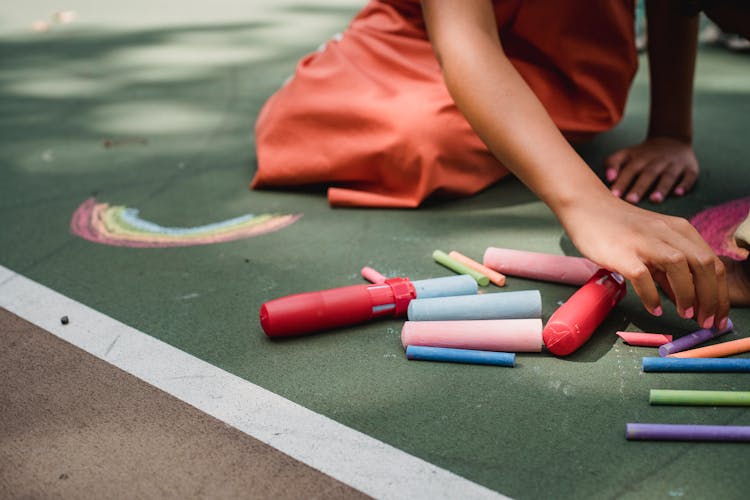 Girl Wearing An Orange Dress Kneeling And Drawing With Chalk On Pavement