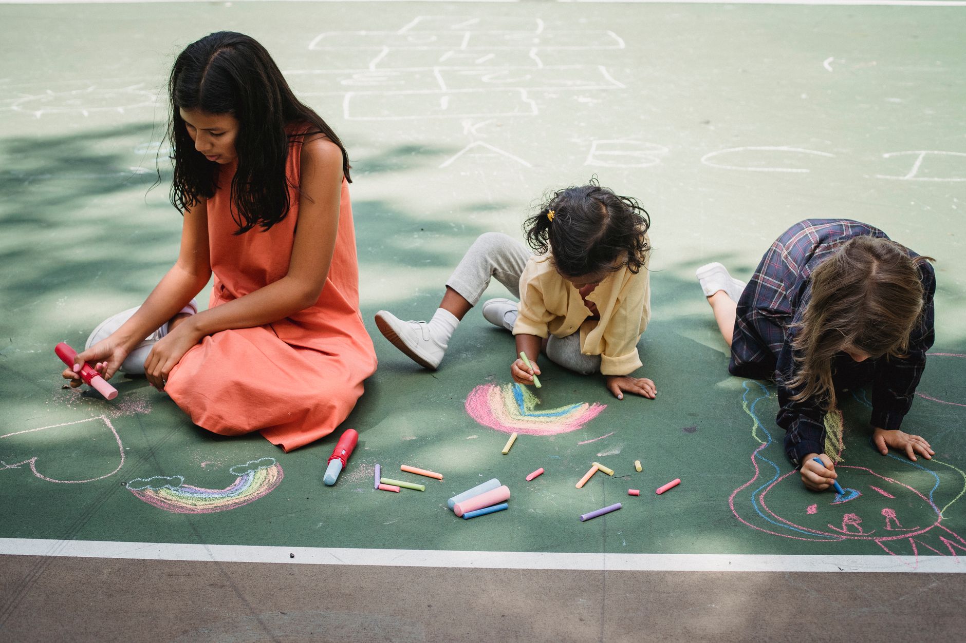 Three girls enjoy a sunny day drawing vibrant chalk art on a playground pavement.