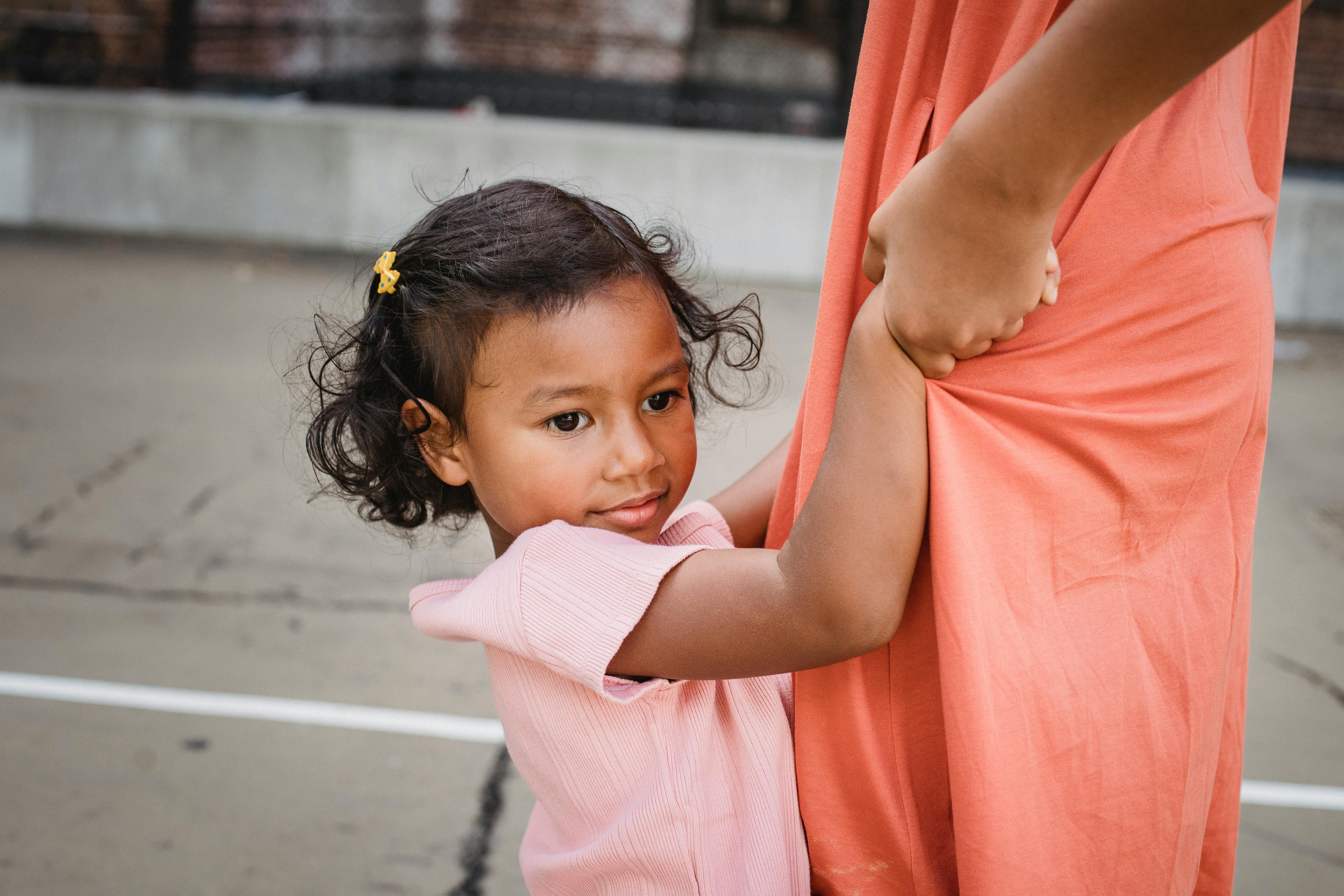 A cute child wearing a pink dress holds onto an adult's dress while outside.