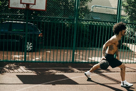 Shirtless man with tattoos performing fitness exercise with medicine ball on outdoor basketball court.