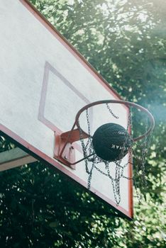 A basketball passing through a chain net on an outdoor hoop surrounded by trees on a sunny day.