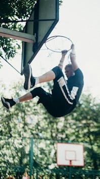 A man performs an energetic dunk, hanging from a basketball hoop outdoors on a bright day.