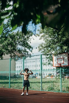 A shirtless man flexes on an outdoor basketball court surrounded by trees and buildings.