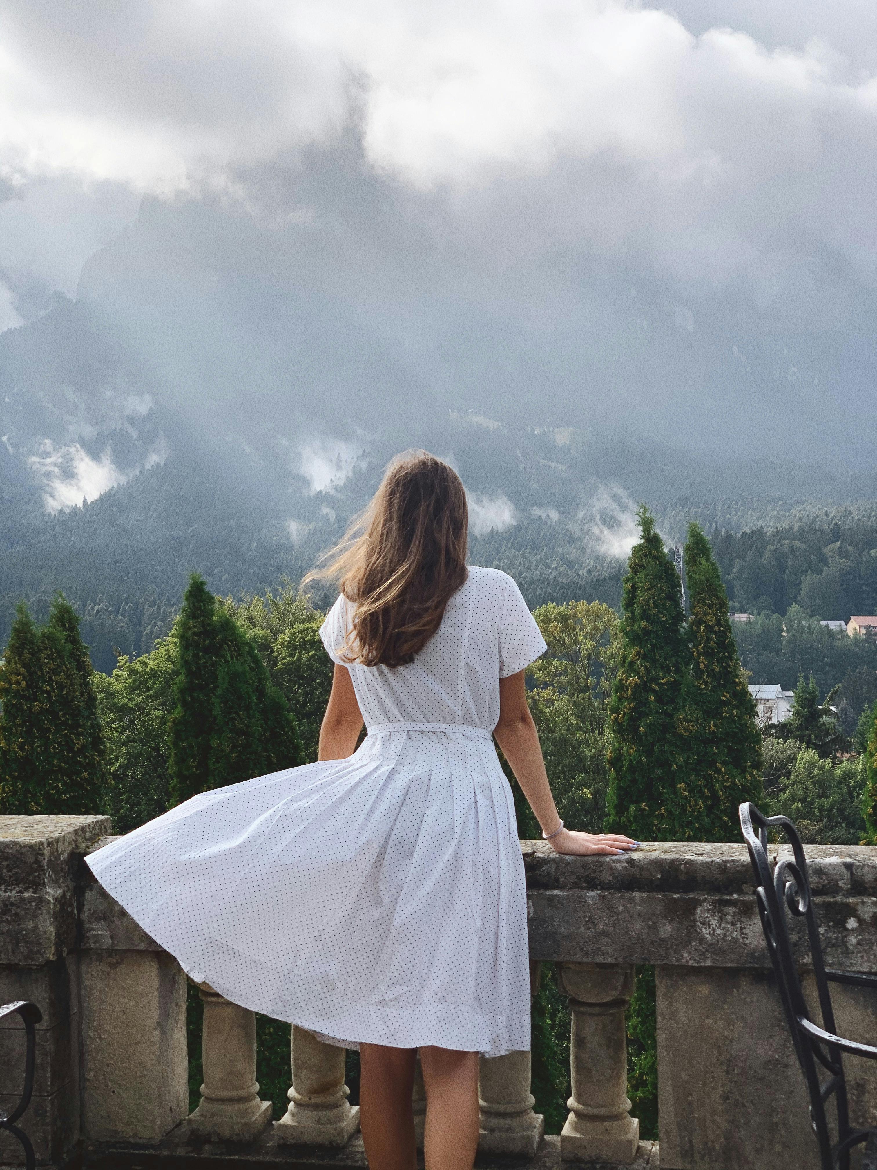 Brunette woman in white dress standing on a balcony overlooking misty mountains and pine trees.