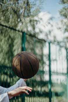Detailed view of a basketball being played outdoors with green fence backdrop, blurred in motion.
