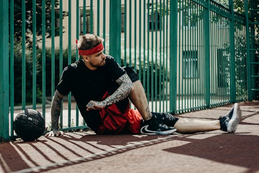 Tattooed basketball player resting on the court beside a green fence.