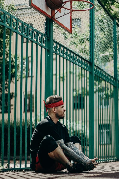 Free Man with tattoos sitting near a basketball court fence under daylight, wearing a red headband. Stock Photo