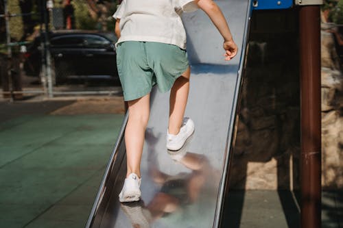 Kid in White Shirt and Green Shorts Climbing Up the Slide in the Playground