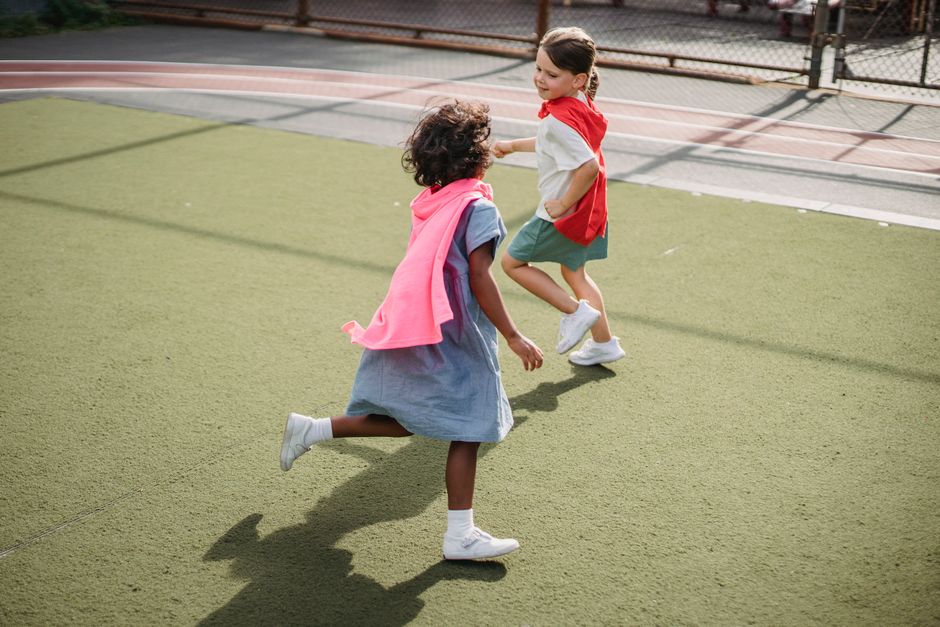 Two children joyfully running with colorful capes on an outdoor playground.