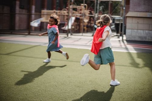 Free Two children enjoying playtime with capes on an outdoor playground, running on a sunny day. Stock Photo