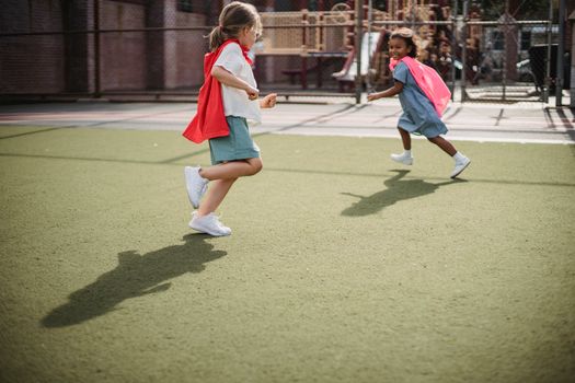 Two young girls play superheroes in a sunny playground, wearing red capes and smiling.