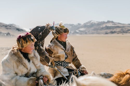 Positive young Mongolian people wearing authentic warm clothes and hats carrying golden eagle on hand and riding horses on vast mountainous valley while looking away contentedly