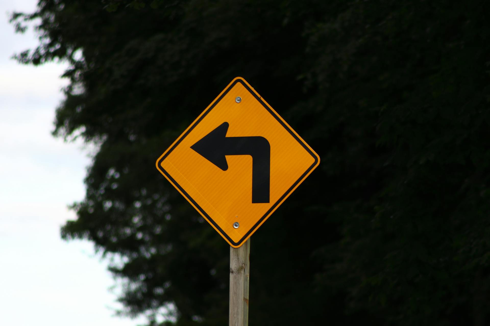 Close-up of a yellow left turn warning sign on a rural road.