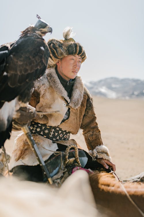 Focused young Mongolian male in authentic warm clothes with eagle on hand riding horse along barren valley and looking away pensively