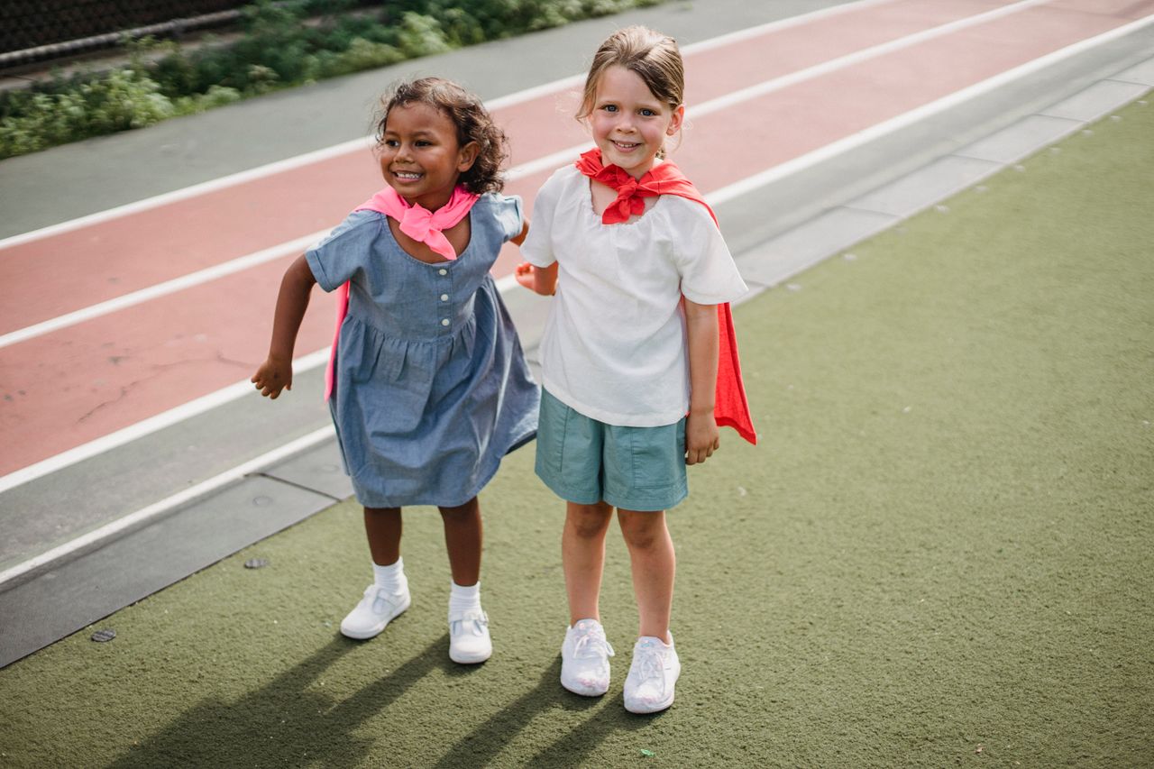 Two young girls wearing capes share a joyful moment on a school playground.