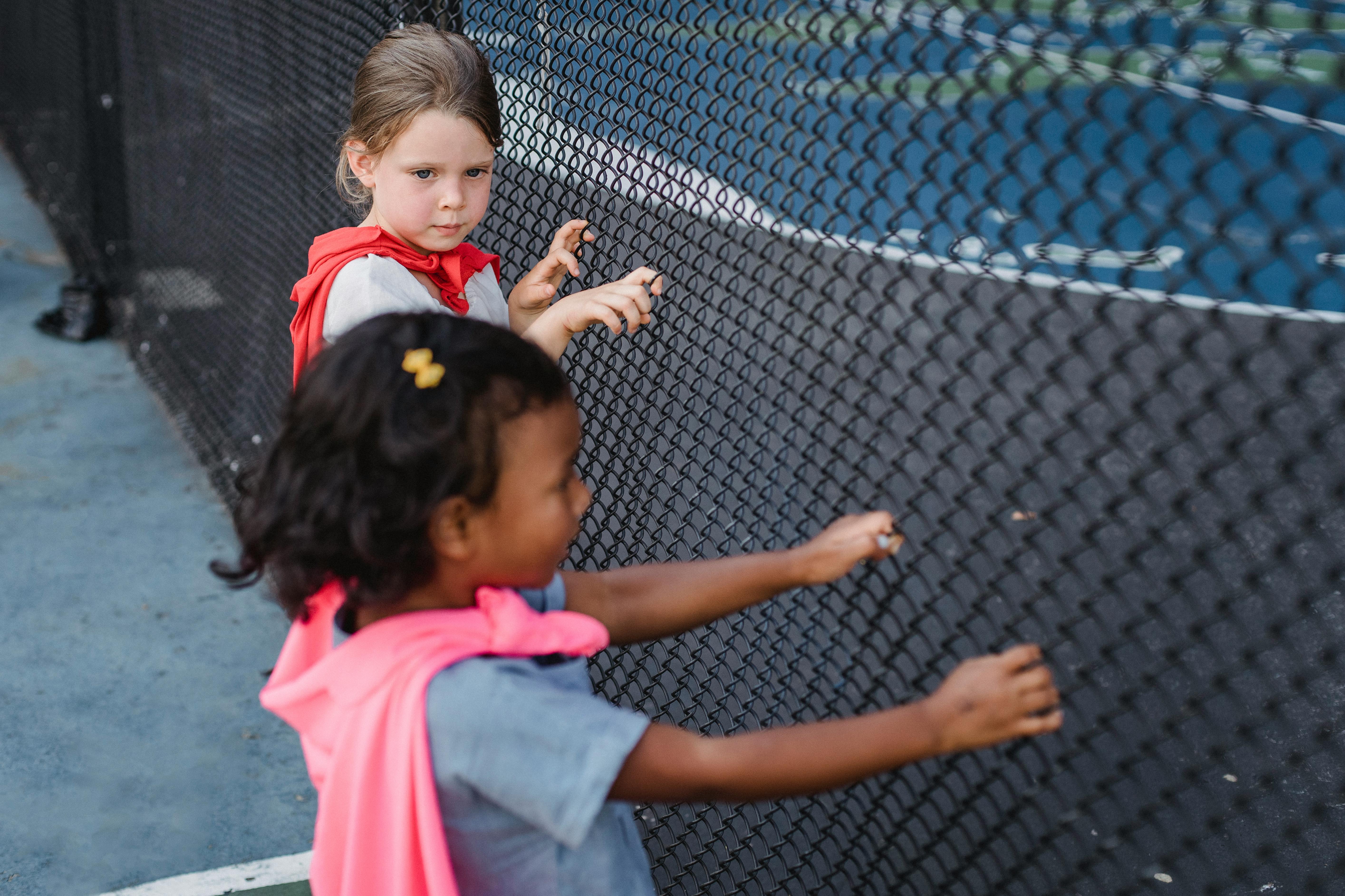 Two young girls peering through a chain-link fence in a playful manner.