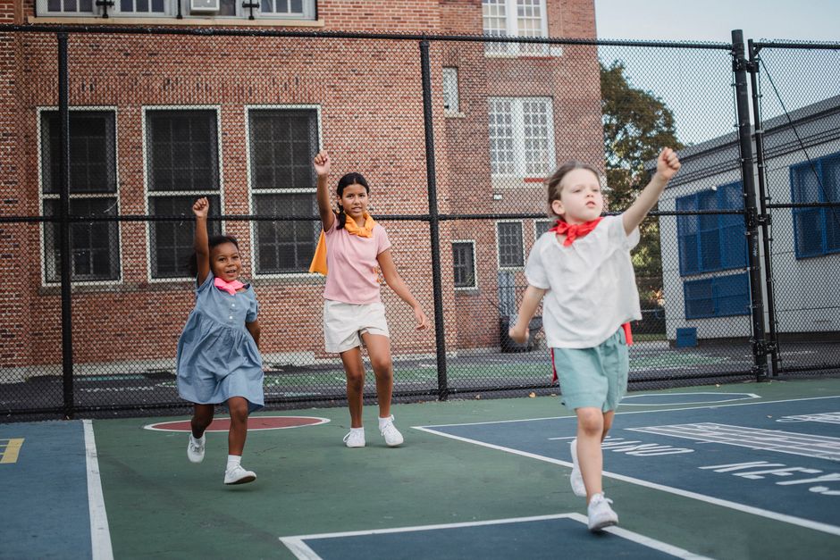 Children role-playing as superheroes during outdoor playtime at a school's basketball court.