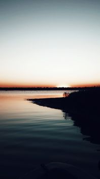 Silhouettes of trees reflecting in rippled river under sky with shiny sun at dusk