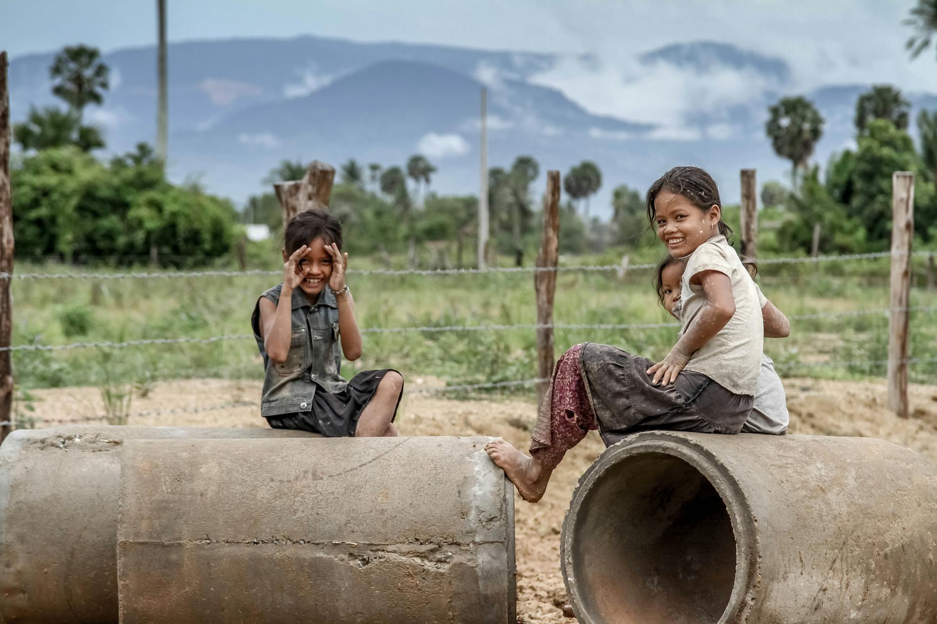 Three children play on concrete pipes in a rural Cambodian landscape with mountains in the background.