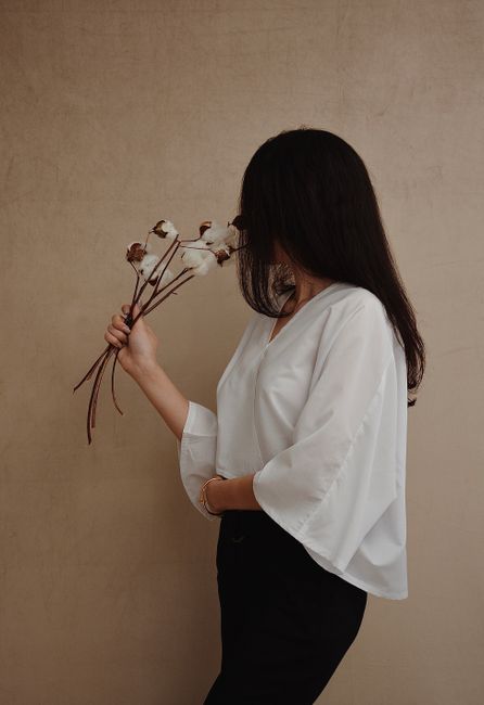 Side view of anonymous lady in stylish white blouse standing with dry plants on beige background