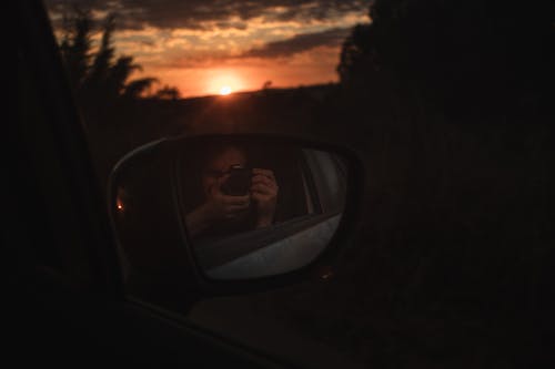 Dark Photograph of a Woman Taking a Selfie in a Side-view Mirror of a Car at Sunset 