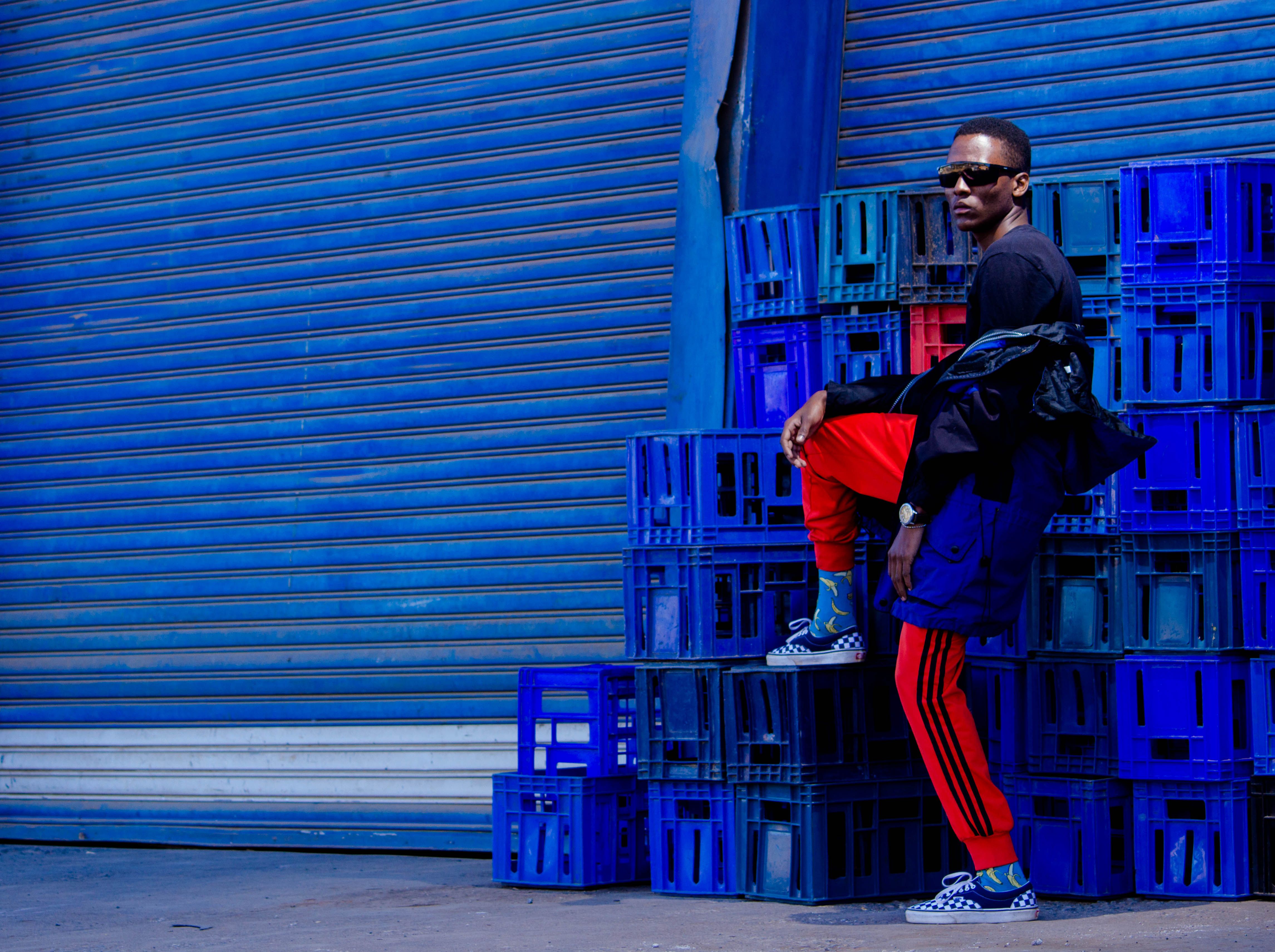 Stylish man posing in vibrant urban setting with colorful crates and shutters.