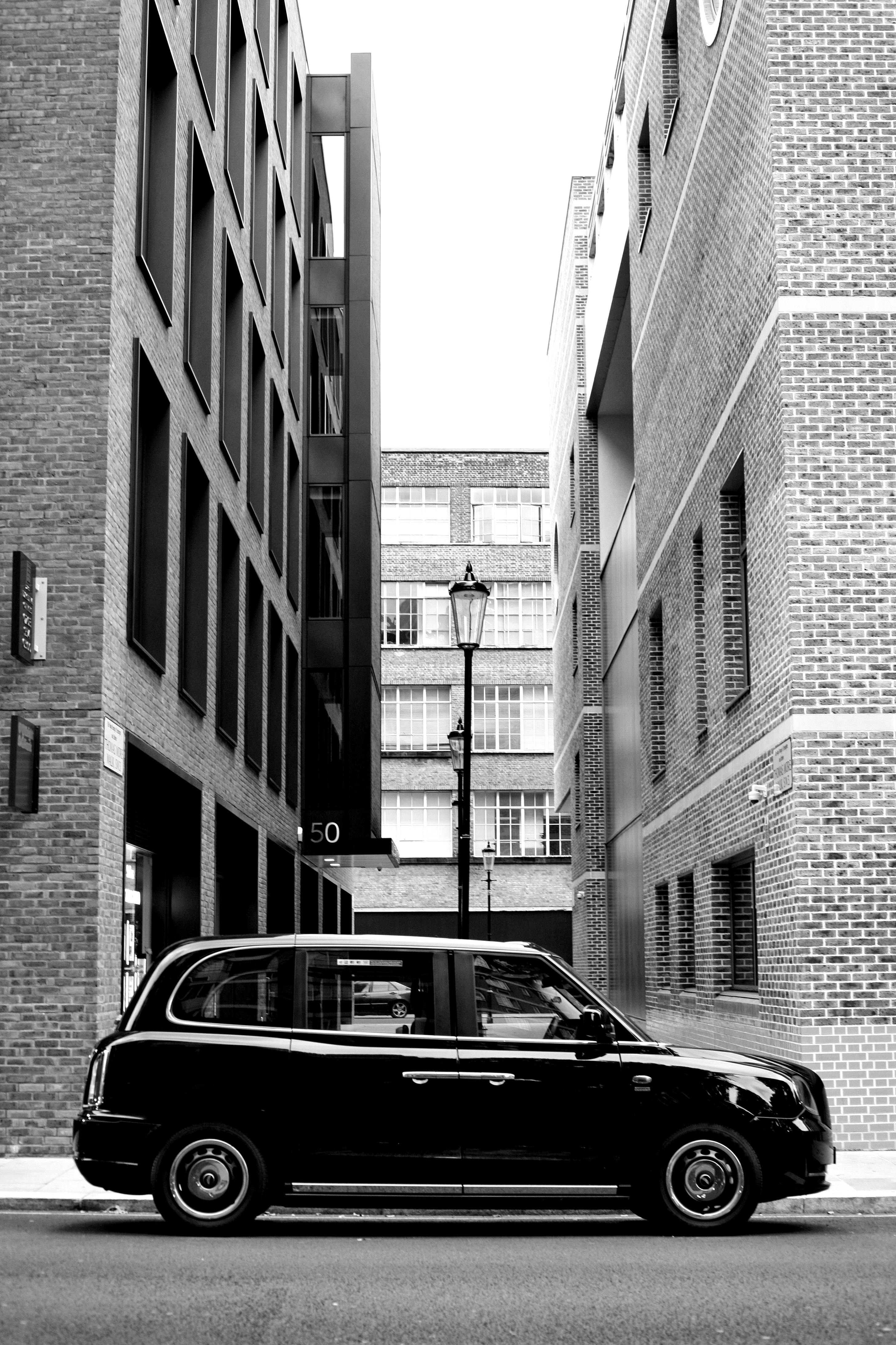 Monochrome photo of a classic car in an urban alleyway between brick buildings.
