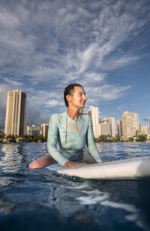 Content young lady in wetsuit floating in sea sitting on surfboard while spending summer holidays in modern resort against blue sky