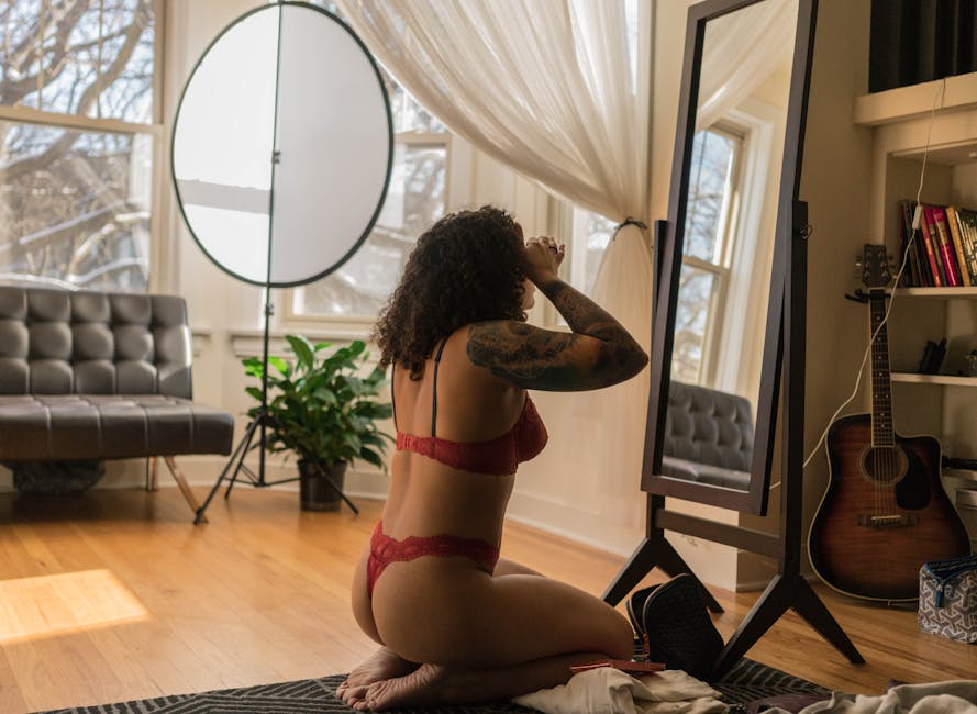 Side view of unrecognizable young sensual tattooed female with curly hair in sexy lingerie sitting on floor in front of mirror and applying makeup