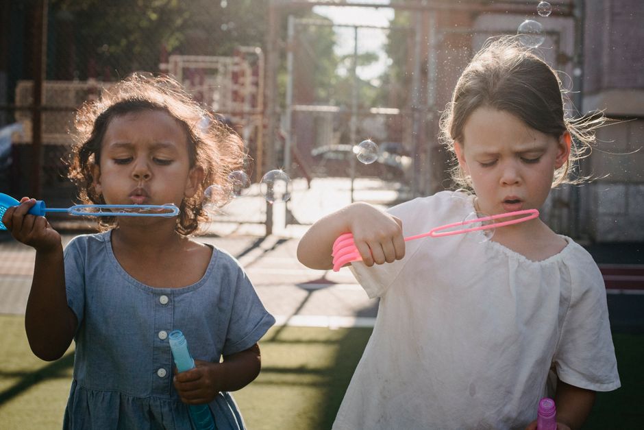 Two young girls enjoy blowing bubbles on a sunny day in a playground.