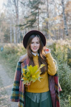 A young woman in a colorful coat and hat holds autumn leaves in a forest setting, embodying fall fashion.