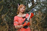 Delighted young female musician in bright pink dress and red gloves with soft cats ears on head playing black ukulele while standing near trees and plants and looking away