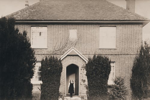 Grayscale Photo Of A Little Girl Standing On The Doorway Of A House With Brickwall