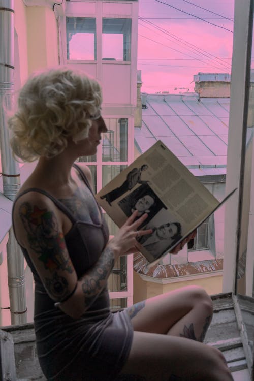 Side view of unrecognizable female with blond hair and tattoos sitting on windowsill with tucked leg near opened window while reading book against pink sky