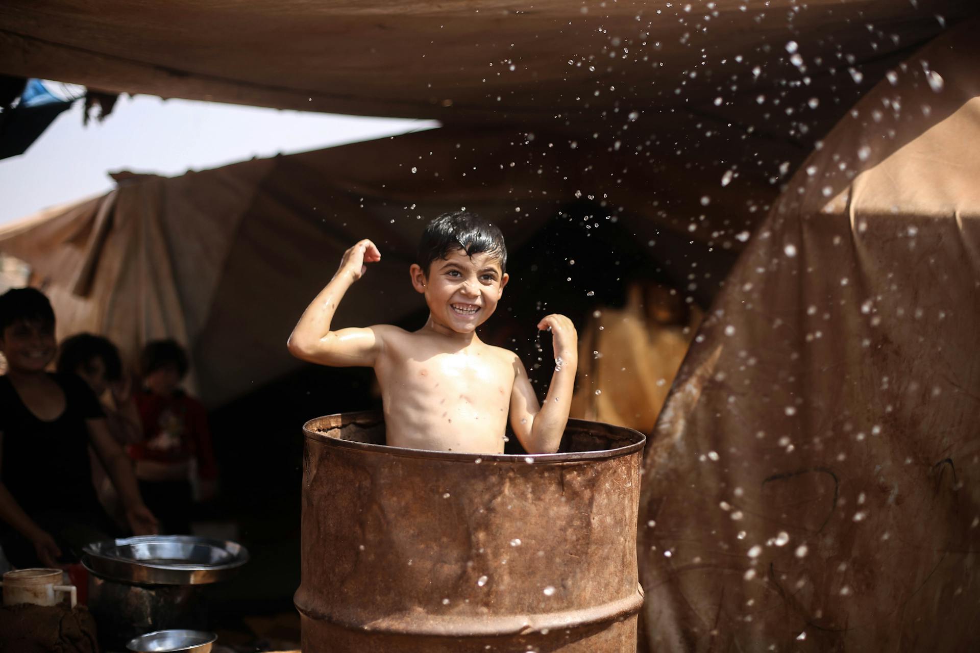 A smiling boy splashes water in a barrel under tents, showcasing joyful moments in a refugee camp.