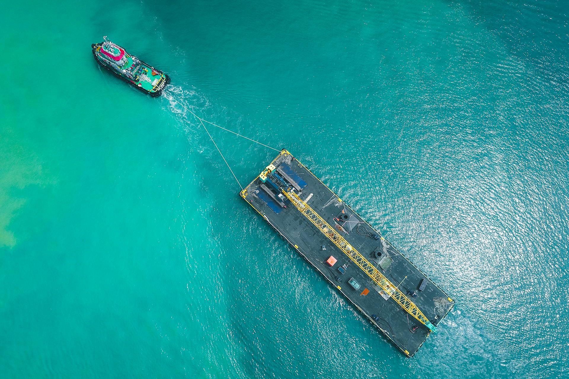 A drone captures a cargo boat towing a large industrial barge across the azure sea.