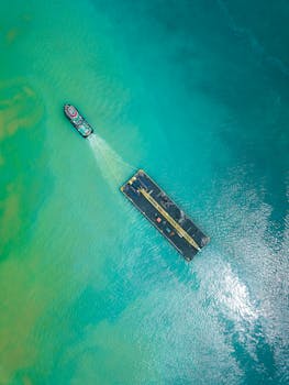Aerial view of a tugboat towing a large barge across a turquoise sea during the day.