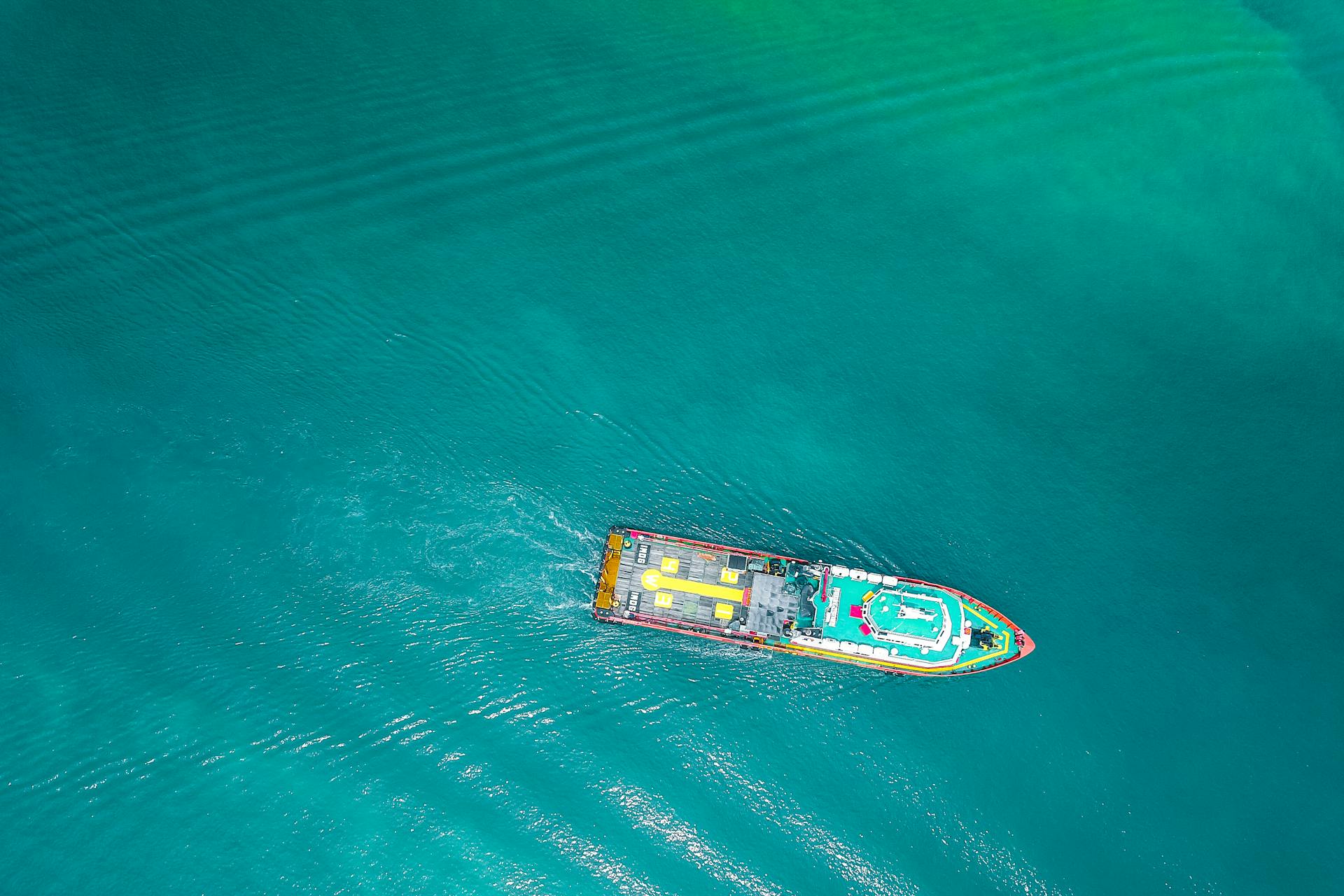Drone view of a cargo ship navigating calm, turquoise waters under clear skies.