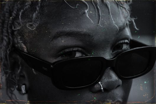 Black and white crop attractive African American female with African hairstyle and nose piercing wearing trendy sunglasses on blurred street and looking at camera attentively