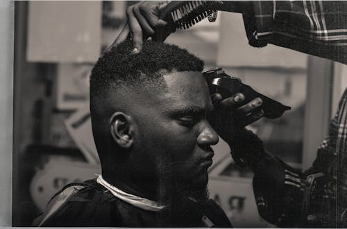 Black and white side view calm adult African American male sitting in barber shop while crop hairdresser trimming hair