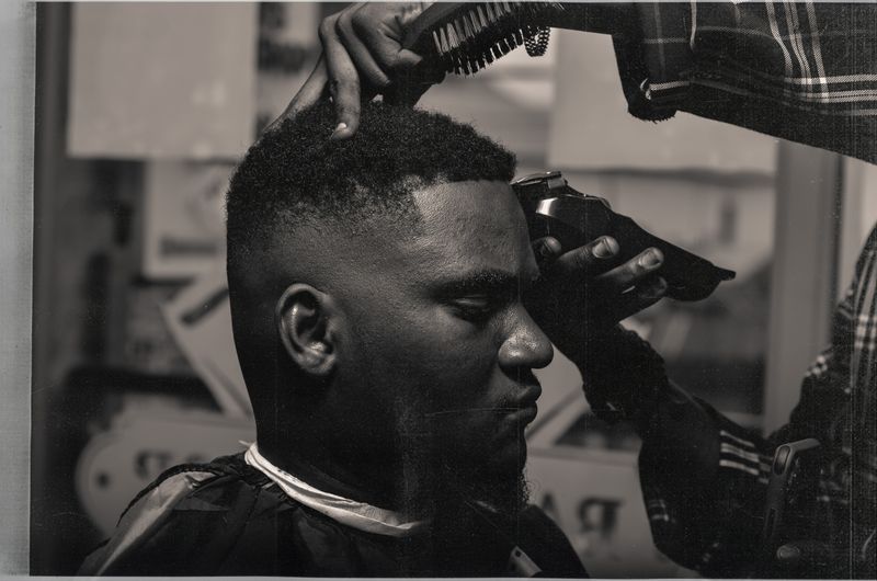 A Black man in a barber cape gets his hairline shaped with clippers at the barbershop, the barber steadying his head with a brush.