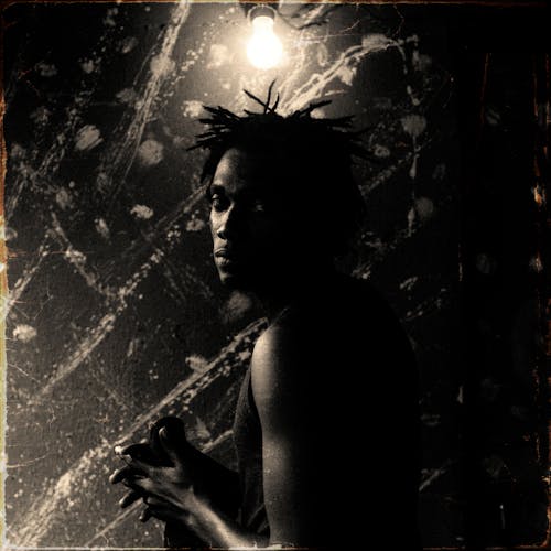 Black and white serious contemplative African American male with dreadlocks wearing casual clothes standing near dark painted wall under electric bulb and looking at camera pensively