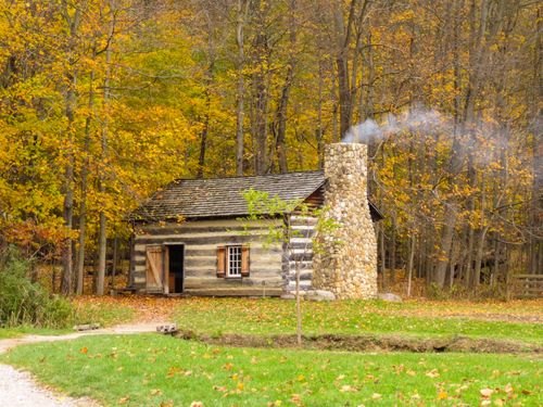 Free Quaint log cabin with stone chimney surrounded by vibrant fall foliage. Stock Photo